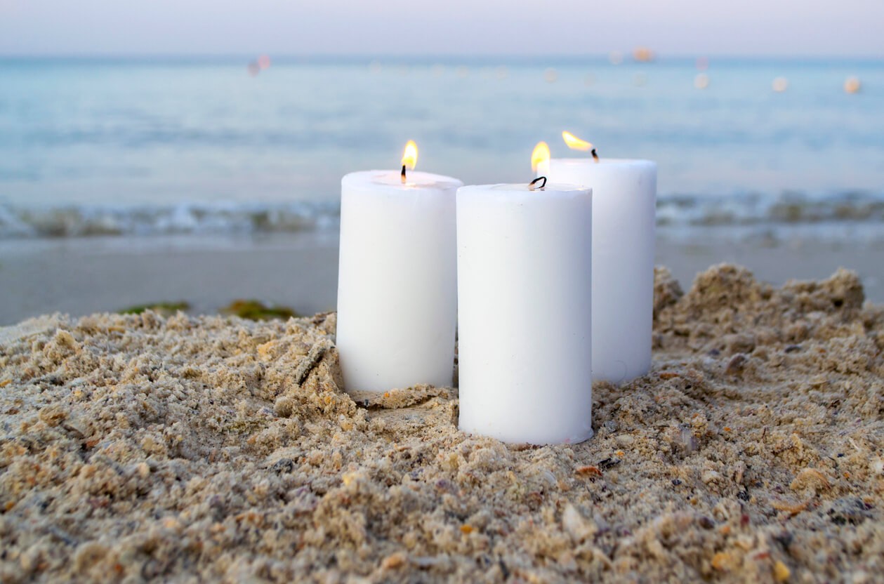 Photo of three white paraffin candles on a beach with the sea in the background. 