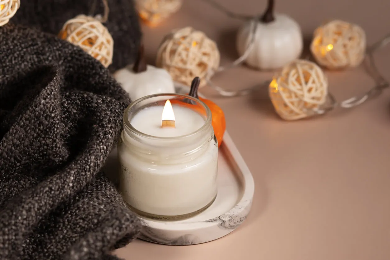 Image of a long-lasting candle made from coconut wax contained in a glass jar, surrounded by a blanket, paper decorations, and small toy pumpkins.